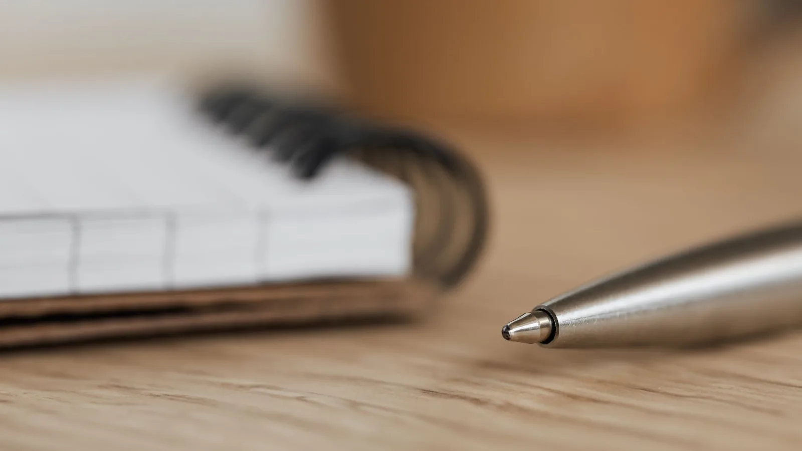A silver ballpoint pen beside a spiral bound notebook on a wooden desk