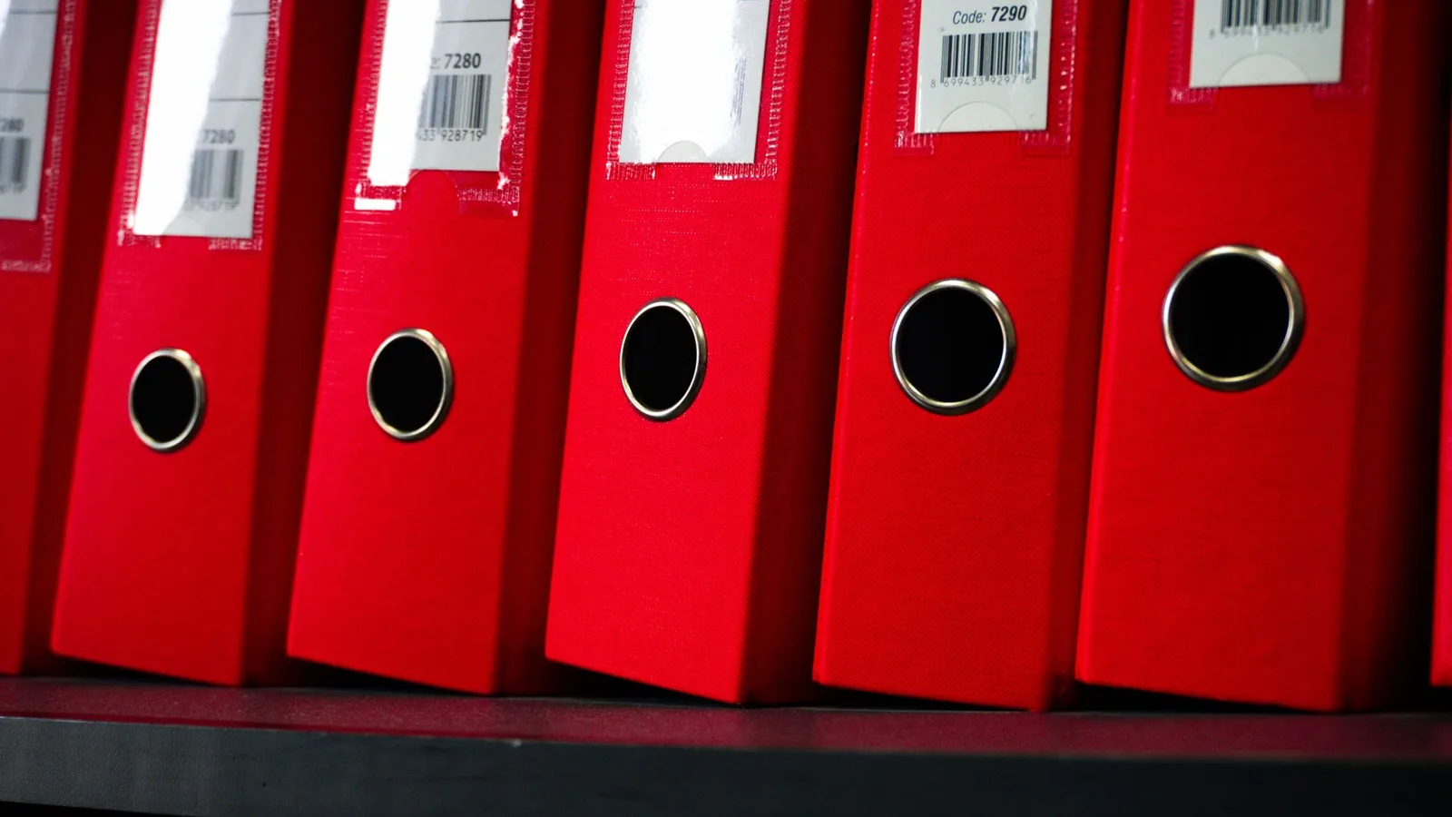 A row of red lever arch files standing on a dark office shelf